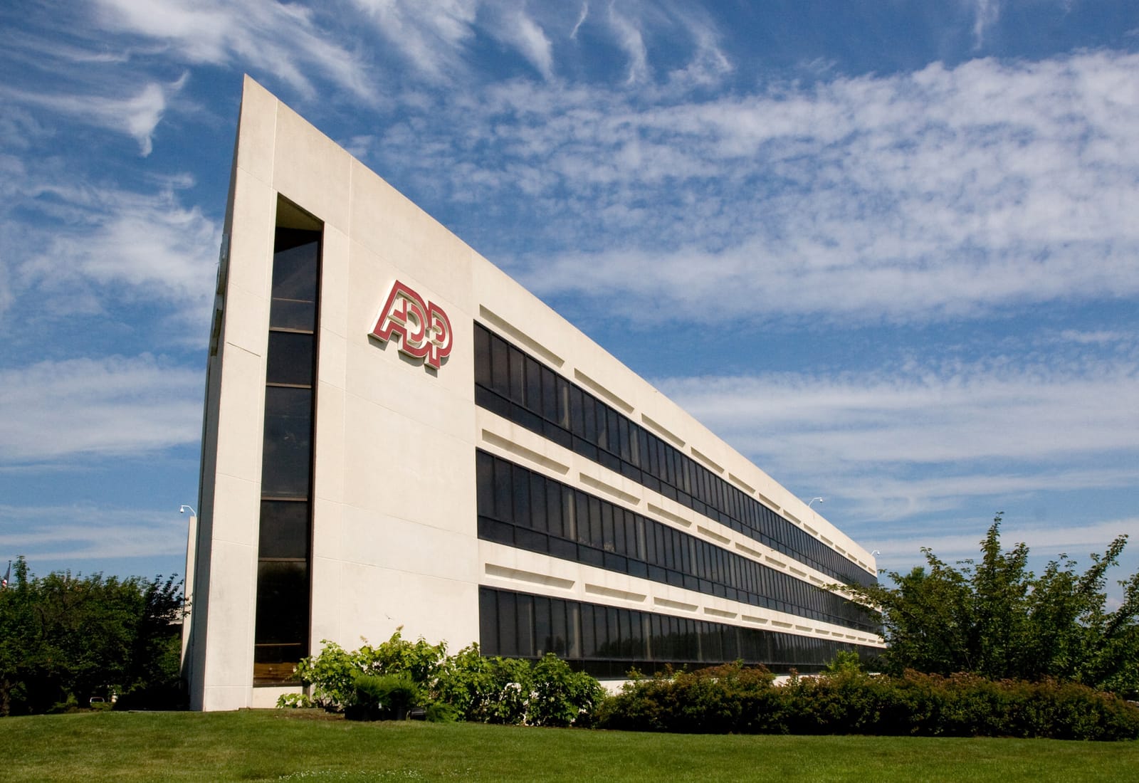 A long, low concrete office building with dark ribbon windows and a red ADP logo on its end wall, seen from the lawn against a blue sky.